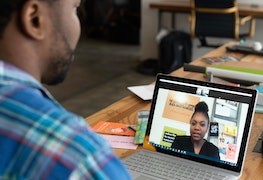 image looking over the shoulder of a man at a desk with a laptop in front of him; the laptop screen shows an image of a woman's head and shoulders, they appear to be video conferencing